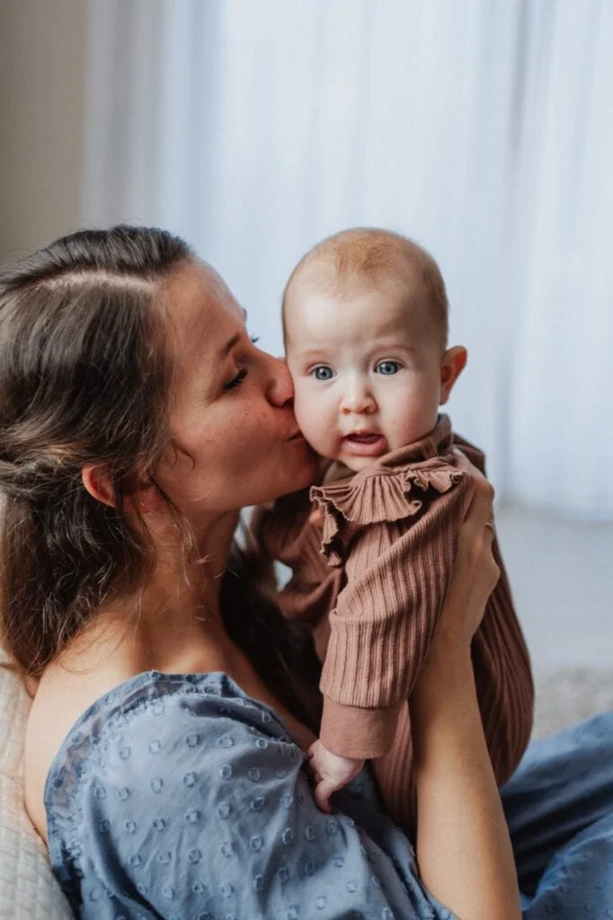 Mutter küsst ihr Baby auf die Wange, helle Vorhänge im Hintergrund