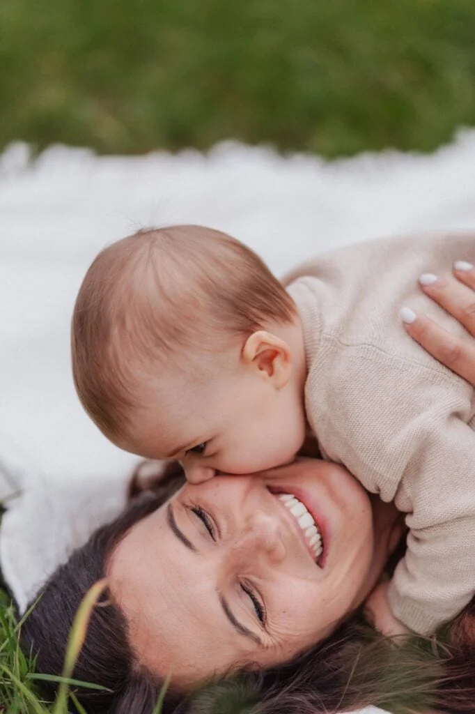 Mutter und Baby liegen lachend im Gras, Baby küsst die Nase der Mutter
