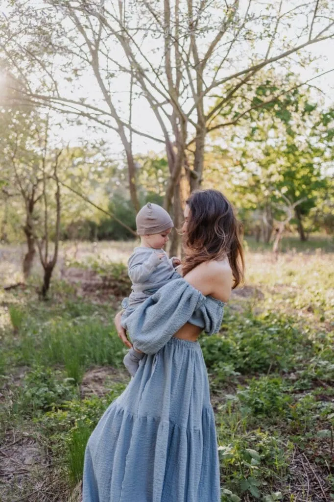 Mutter im blauen Leinenkleid trägt ihr Kind über einen Feldweg im Abendlicht