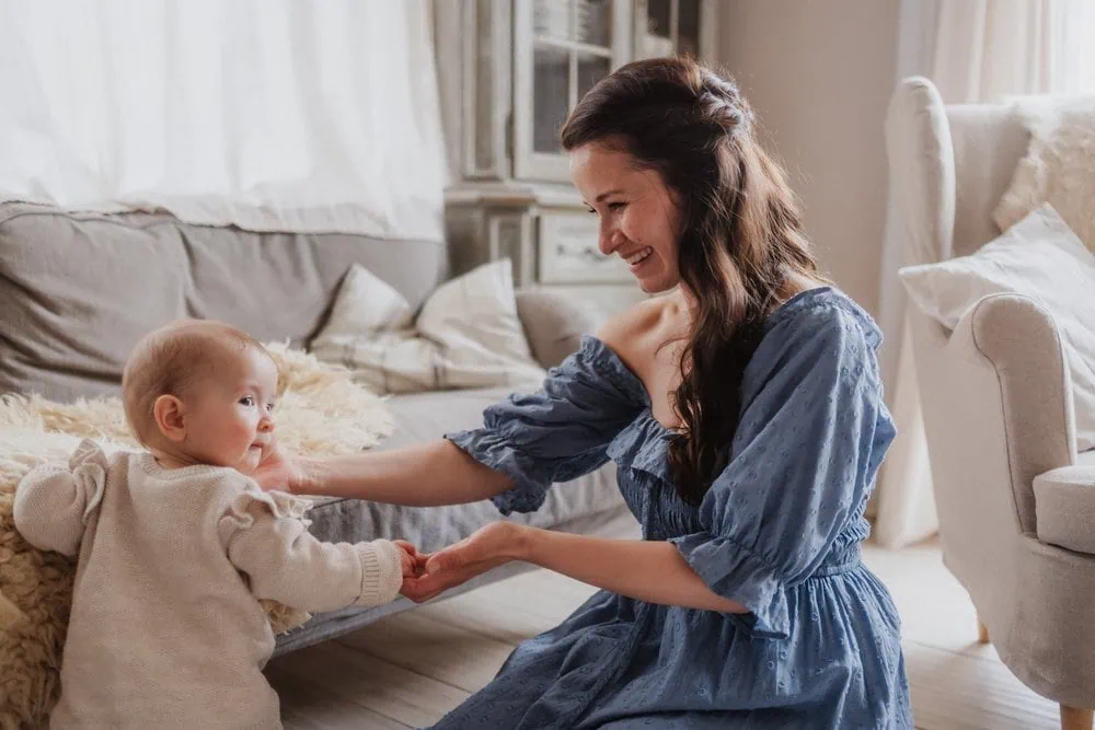 Mutter im blauen Kleid hält lachend die Hände ihres Babys in einem hellen Wohnzimmer